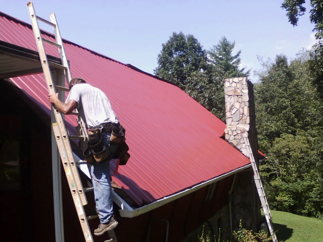 Roofer on a ladder beside a red metal roof and stone chimney — Soffit & Fascia Repair by Roofing Brothers in Gulf
